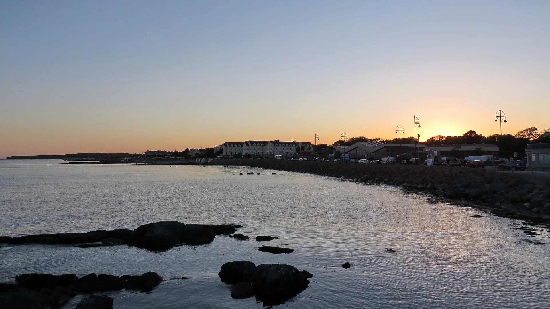 The Salthill seafront at sunset, with promenade lamp-posts and low buildings silhouetted beyond a rock seawall and the calm water of Galway Bay