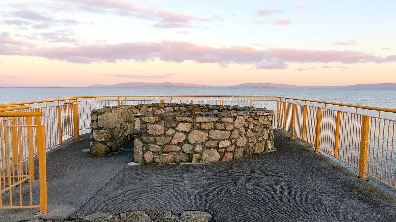 A stone-walled viewpoint behind yellow railings on the Salthill promenade, with Galway Bay and the hills of County Clare under a pink dusk sky